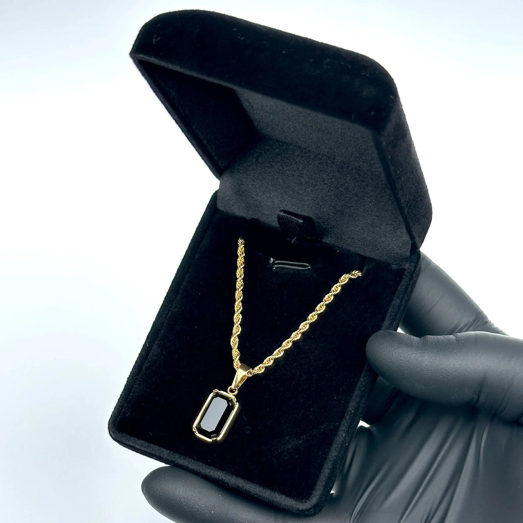 Gold necklace with a black pendant in an open black jewelry box held by a gloved hand.