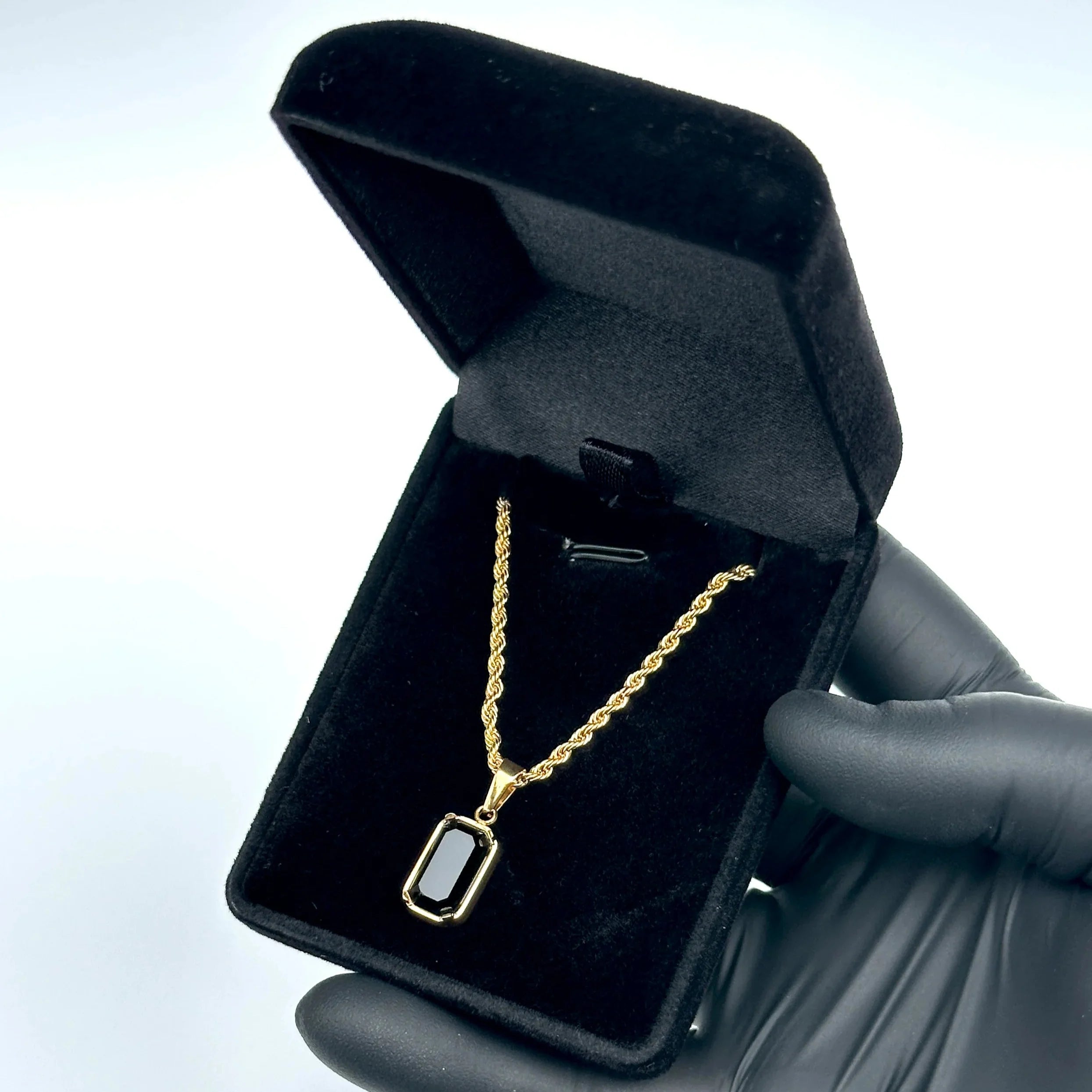 Gold necklace with a black pendant in an open black jewelry box held by a gloved hand.